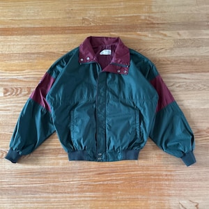 May include: A dark green windbreaker jacket featuring a deep burgundy collar and sleeve panels. The jacket includes a front zipper closure, two side pockets, and dark green ribbed cuffs and hem. A white label is visible inside the collar.
