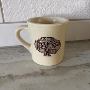 May include: A cream-coloured ceramic mug with a speckled finish and a single handle. The front features a brown graphic logo with the text Jack Daniel's Tennessee Mud in a stylised font. The mug has a smooth, rounded rim and a sturdy base.