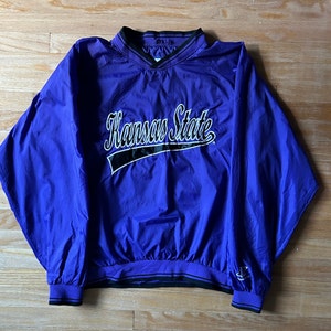 May include: A purple windbreaker jacket with the text "Kansas State" in white and black lettering. The jacket has a black zipper and black trim around the collar, cuffs, and hem.