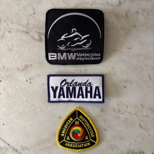 May include: Three embroidered patches. The top patch is black with a white motorcycle graphic and the text "BMW Motorcycles of Daytona Beach." The middle patch is white with "Orlando YAMAHA" in blue. The bottom patch is yellow with "AMERICAN MOTORCYCLE ASSOCIATION."