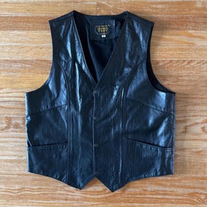 May include: A black leather waistcoat featuring a V-neckline, four front snap button closures, and two side pockets. The interior label displays the text VETO CANADA. The garment is constructed with visible seams and a smooth, dark finish throughout.