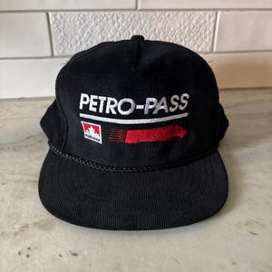May include: A black corduroy baseball cap featuring the white embroidered text PETRO-PASS above a small red and white Canadian maple leaf logo and a stylised red truck graphic. The cap includes a black braided cord trim across the base of the crown.