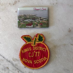 May include: A rectangular magnet featuring a photographic landscape of Peggy's Cove, Nova Scotia, and a circular embroidered patch in red with yellow trim, green leaves, and the text KINGS DISTRICT CJ'77 NOVA SCOTIA stitched in yellow thread.