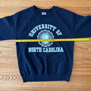 Op de afbeelding: Marineblauw sweatshirt met de tekst "UNIVERSITY OF NORTH CAROLINA" in wit en lichtblauw. Het sweatshirt wordt gemeten met een gele meetlint, met een lengte van ongeveer 61 cm.