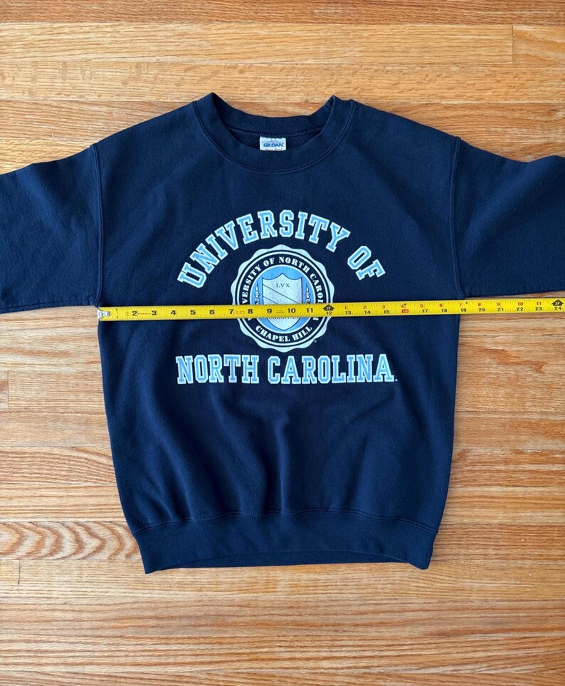 Op de afbeelding: Marineblauw sweatshirt met de tekst "UNIVERSITY OF NORTH CAROLINA" in wit en lichtblauw. Het sweatshirt wordt gemeten met een gele meetlint, met een lengte van ongeveer 61 cm.