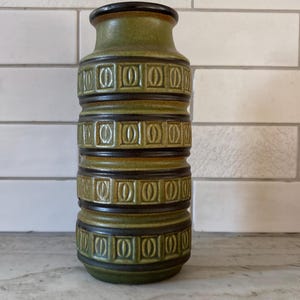 May include: A green ceramic vase with a cylindrical shape and a dark brown rim. The vase features a repeating pattern of rectangular shapes with rounded corners, separated by dark brown bands. The vase is approximately 25 cm tall.