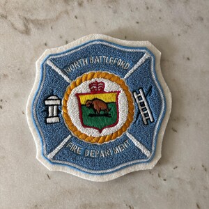 May include: Embroidered patch featuring the North Battleford Fire Department emblem. The patch has a light blue background with a white border. The center emblem includes a beaver, crown, and ladder.