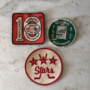 May include: Three vintage patches. One red square patch with "Winnipeg Junior A&W Winter Games" text. A green circular patch with "Regional Int. Silver Stick 1979 Bloomington" text. A circular patch with "Stars" text and hockey sticks.
