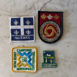 May include: Four embroidered patches including a square blue Quebec patch with white fleur-de-lis, a shield-shaped Canada Fitness Award patch, a square 1974 Scout Camporee Winnipeg patch with yellow and blue, and a small West Van patch with a lighthouse.
