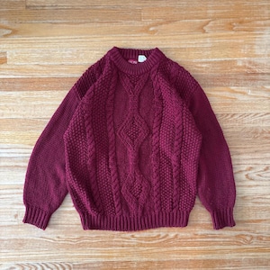 May include: A burgundy cable-knit sweater with a crew neck and ribbed cuffs and hem. The sweater features a central diamond pattern and textured knit details. The garment is made of a thick, warm material, perfect for cooler weather.