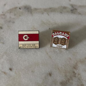 Könnte beinhalten: Zwei rechteckige Metallanstecknadeln von den Olympischen Winterspielen 1988 in Calgary. Die linke Anstecknadel zeigt ein rot-weißes Flaggendesign mit Logo und dem Text HOST CITY 1988 OLYMPIC WINTER GAMES. Die rechte Anstecknadel zeigt CALGARY, 1988 und Ringe.