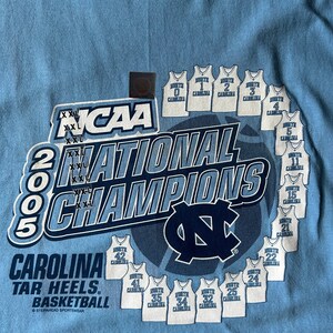 May include: A light blue t-shirt with a white graphic of the 2005 NCAA National Champions, the University of North Carolina Tar Heels basketball team. The graphic includes the team's logo, the year 2005, and the text "NCAA National Champions". The graphic also includes the jersey numbers of the players on the team.