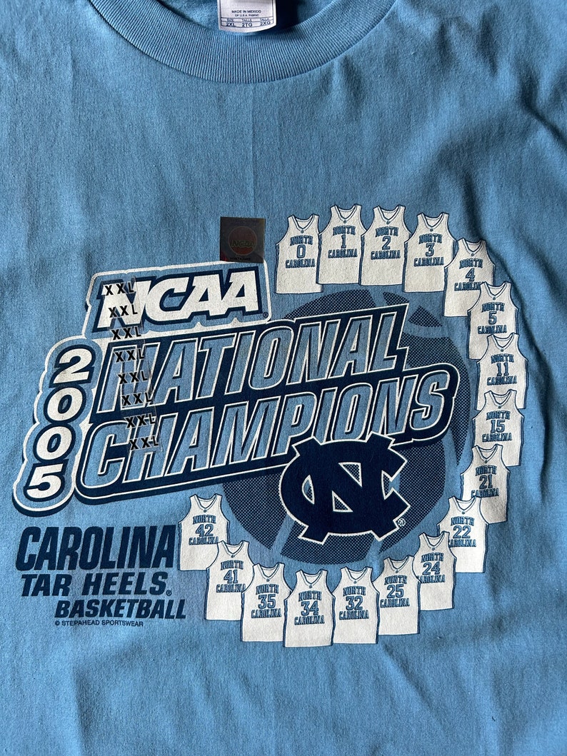May include: A light blue t-shirt with a white graphic of the 2005 NCAA National Champions, the University of North Carolina Tar Heels basketball team. The graphic includes the team's logo, the year 2005, and the text "NCAA National Champions". The graphic also includes the jersey numbers of the players on the team.