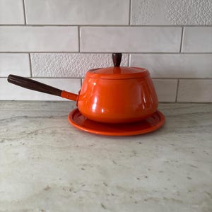 Può includere: Una casseruola arancione smaltata con coperchio e manico in legno. La casseruola è appoggiata su un piatto arancione abbinato. Il manico e il pomello del coperchio sono in legno scuro. Lo sfondo è una parete di piastrelle bianche.