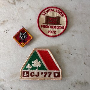 Könnte beinhalten: Drei Vintage-Stickabzeichen. Eines ist eine rautenförmige Form in Rot und Gelb mit einer Wolfsgrafik und dem Wort "WOLF". Ein runder Aufnäher lautet "St. Vital Cubs Frontier Days 1972". Ein dritter Aufnäher ist grün, rot und weiß mit "CJ '77".