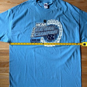 May include: A light blue t-shirt with a graphic of the 2005 NCAA National Champions, the Carolina Tar Heels. The graphic features the team's logo and a basketball with the number 2005.
