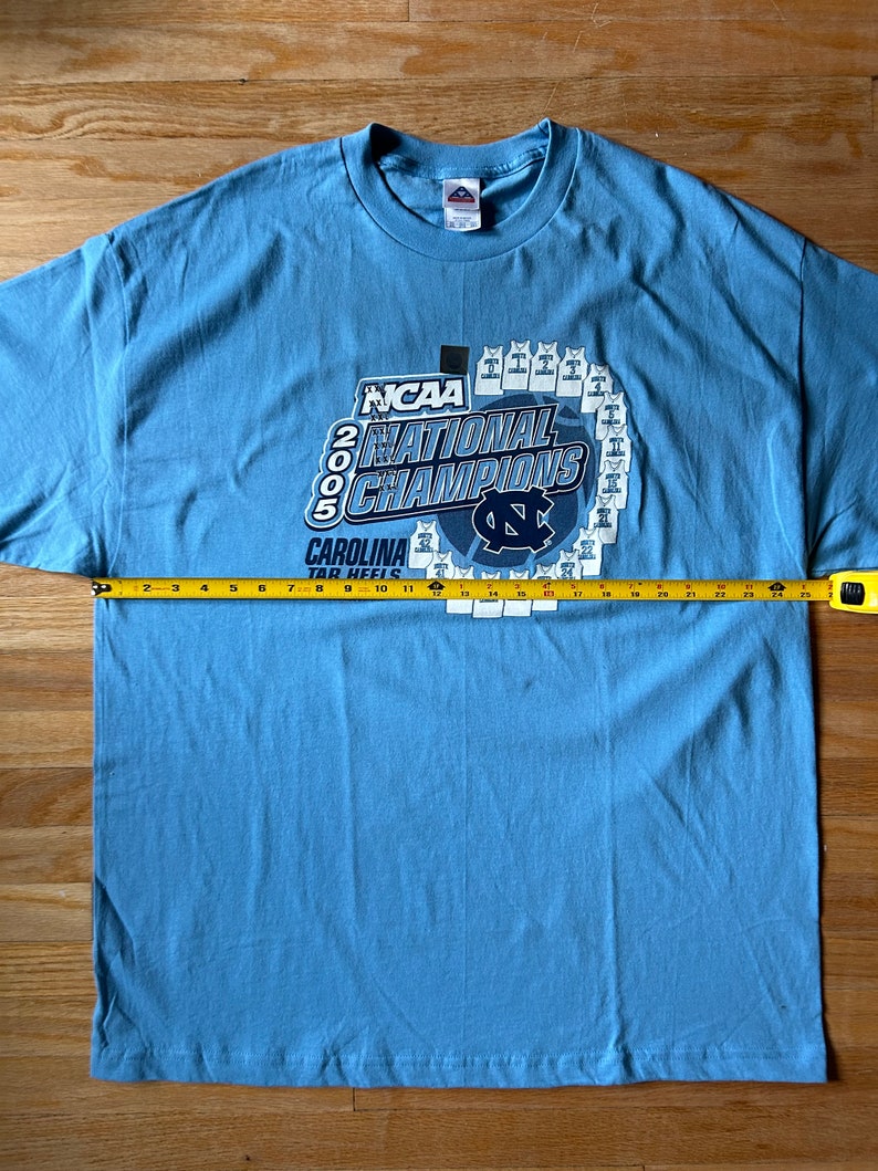 May include: A light blue t-shirt with a graphic of the 2005 NCAA National Champions, the Carolina Tar Heels. The graphic features the team's logo and a basketball with the number 2005.