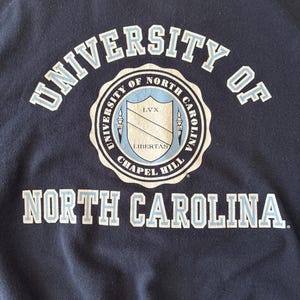 Op de afbeelding: Marineblauwe sweatshirt met de tekst "UNIVERSITY OF NORTH CAROLINA" in witte en lichtblauwe letters. Een cirkelvormig embleem in het midden toont een schild en de woorden "CHAPEL HILL" en "LIBERTAS".