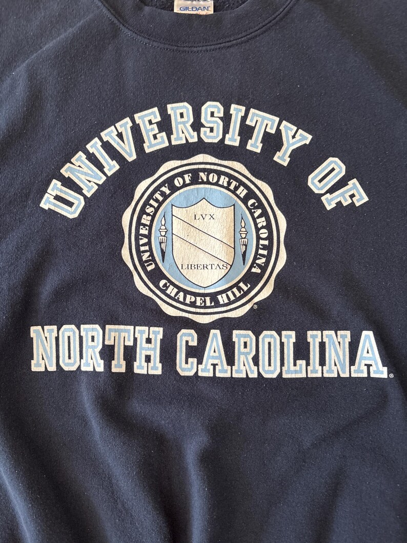 Op de afbeelding: Marineblauwe sweatshirt met de tekst "UNIVERSITY OF NORTH CAROLINA" in witte en lichtblauwe letters. Een cirkelvormig embleem in het midden toont een schild en de woorden "CHAPEL HILL" en "LIBERTAS".