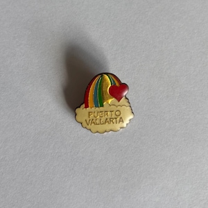Può includere: Una piccola spilla dorata con un design arcobaleno con un cuore rosso e le parole "PUERTO VALLARTA". L'arcobaleno ha strisce gialle, arancioni, verdi e blu. La spilla ha la forma di una nuvola.