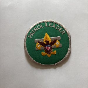 May include: A circular green embroidered patch with the words "PATROL LEADER" in white. The patch features a gold fleur-de-lis and a brown emblem with red, white, and blue accents. The patch has a silver border.