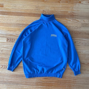 May include: Bright blue turtleneck sweatshirt with long sleeves. The word "BOCA" is embroidered in white above the chest. The sweatshirt is made of a soft material and is laid flat on a wooden surface.
