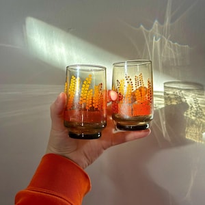 May include: Two clear glass tumblers with an orange and yellow floral design. The glasses are being held in a hand.