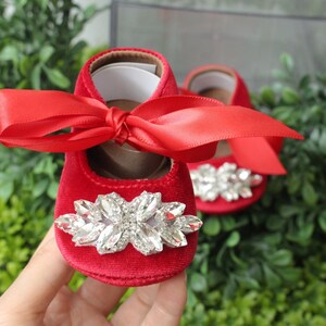 Peut inclure: Chaussures de b&eacute;b&eacute; en velours rouge avec un grand embellissement en strass et un n&oelig;ud en satin rouge.