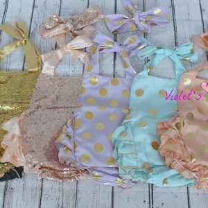 Pode incluir: Cinco conjuntos para menina com detalhes dourados. Os conjuntos incluem um macacão de lantejoulas douradas, um macacão de lantejoulas cor-de-rosa, um macacão de bolinhas lavanda, um macacão de bolinhas azul claro e um macacão de bolinhas pêssego. Cada conjunto vem com uma faixa de cabelo combinando.