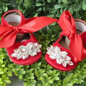 Peut inclure: Une paire de chaussures de b&eacute;b&eacute; en velours rouge avec un grand embellissement en strass et un n&oelig;ud en satin rouge.
