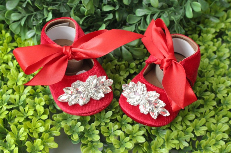 Peut inclure: Une paire de chaussures de b&eacute;b&eacute; en velours rouge avec un grand embellissement en strass et un n&oelig;ud en satin rouge.