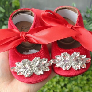 Peut inclure: Une paire de chaussures de b&eacute;b&eacute; en velours rouge avec un grand embellissement en strass sur l'orteil et un n&oelig;ud en satin rouge sur le dessus. Les chaussures sont sur une main.