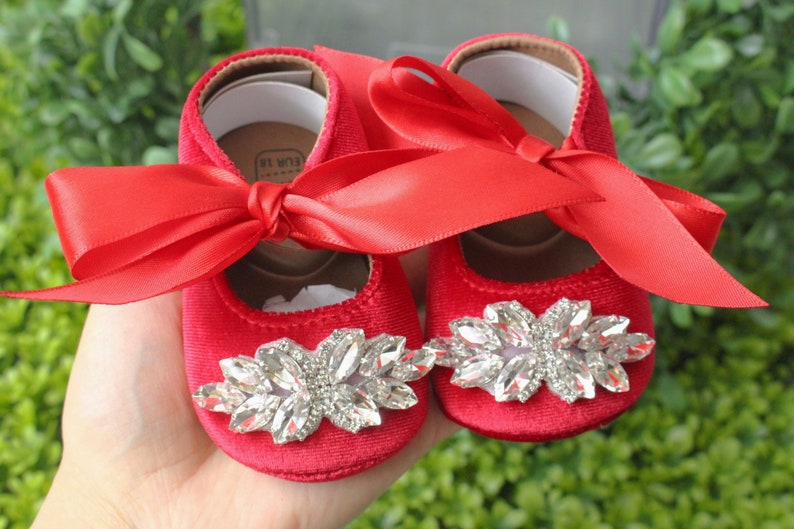 Peut inclure: Une paire de chaussures de b&eacute;b&eacute; en velours rouge avec un grand embellissement en strass sur l'orteil et un n&oelig;ud en satin rouge sur le dessus. Les chaussures sont sur une main.