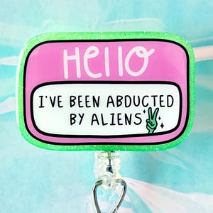 May include: A badge reel with a pink and green design. The badge has the word "HELLO" in white, and the text "I'VE BEEN ABDUCTED BY ALIENS" with a peace sign. The reel has a clear plastic strap and a metal clip.