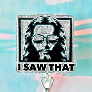 May include: A rectangular badge reel with a silver glitter border and a black and white image of a man wearing sunglasses. The text "I SAW THAT" is below the image. The background is a mix of blue, pink, and white.
