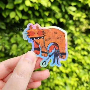 Könnte beinhalten: Ein holografischer Aufkleber mit einer orangefarbenen Cartoon-Katze mit einem Stachelhalsband und einer Kettenleine, die von einer kleinen Figur gehalten wird. Die Katze hat schwarze Akzente, vor einem verschwommenen grünen Hintergrund.