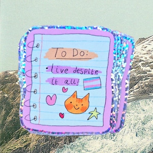Könnte beinhalten: Ein notizbuchförmiger Aufkleber mit einer hellblauen, linierten Seite und einem lila Rand. Auf der Seite steht der Text "To Do: live despite it all!", zusammen mit einem Katzenkopf, einem Herzen, einem Stern und einer Transgender-Pride-Flagge.