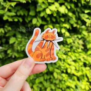 Könnte beinhalten: Ein Aufkleber mit einer orangefarbenen Katze mit einem stacheligen Halsband. Die Katze hat ein lächelndes Gesicht und ist mit dunkelorangefarbenen Streifen und Punkten verziert. Der Aufkleber wird vor einem grünen, blättrigen Hintergrund gehalten.