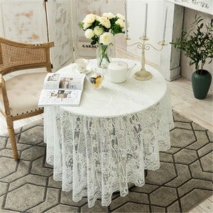May include: White lace tablecloth with a delicate floral pattern, draped over a round table with a white vase of white flowers, a book, and a teapot. The table is set in a room with white walls and a wooden chair.