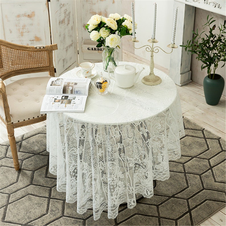 May include: White lace tablecloth with a delicate floral pattern, draped over a round table with a white vase of white flowers, a book, and a teapot. The table is set in a room with white walls and a wooden chair.