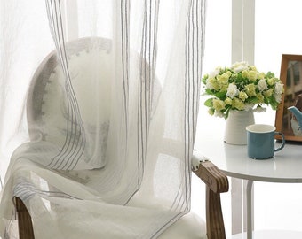 May include: Sheer white curtains with vertical gray stripes hang in front of a white chair. A small white table holds a vase of flowers, a blue mug, and a framed photo, creating a bright, airy interior scene.