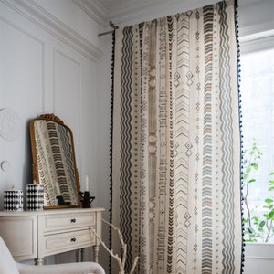 Puede incluir: Un par de cortinas beige con un patrón geométrico en blanco y negro. Las cortinas tienen borlas negras a lo largo de los bordes.