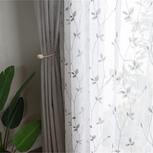 May include: Grey and white sheer curtains with a leaf pattern. The curtains are held together with a wooden curtain tieback.
