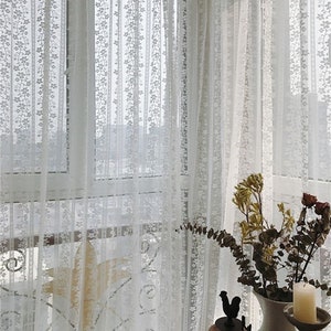 Puede incluir: Cortinas de encaje blanco con un patr&oacute;n floral cuelgan en una ventana. Las cortinas son transparentes y permiten que la luz pase a trav&eacute;s de ellas.