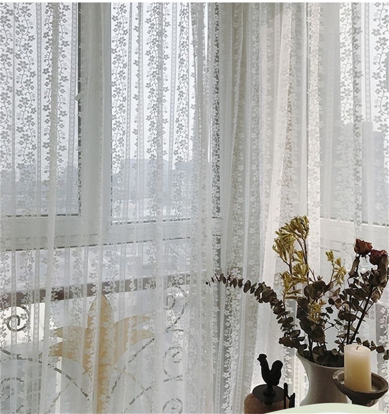 Puede incluir: Cortinas de encaje blanco con un patr&oacute;n floral cuelgan en una ventana. Las cortinas son transparentes y permiten que la luz pase a trav&eacute;s de ellas.