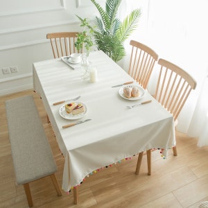 Pode incluir: Toalha de mesa branca com uma borda de pompons coloridos, colocada em uma mesa de madeira com duas cadeiras e um banco. A mesa é decorada com um prato de doces e um prato de biscoitos.