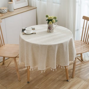 May include: A round, beige linen tablecloth with a fringed border. The tablecloth is draped over a wooden table with a vase of flowers in the centre. Two wooden chairs are visible in the background.