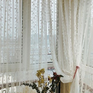 Puede incluir: Cortinas blancas de encaje con un patr&oacute;n floral cuelgan en una ventana. Las cortinas est&aacute;n atadas con una cinta rosa.