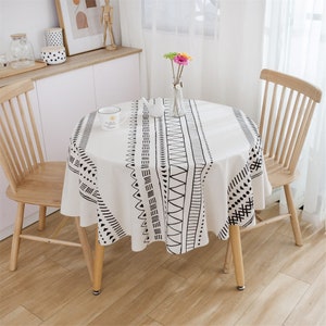 May include: A round white tablecloth with a black geometric pattern. The tablecloth is on a round wooden table with two wooden chairs.
