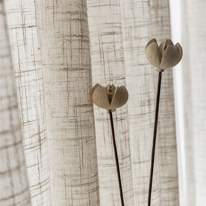 Puede incluir: Dos tallos de flores secas con cabezas de flores beige sobre un fondo de cortinas de lino blanco transparentes con un sutil patrón de cuadrícula.